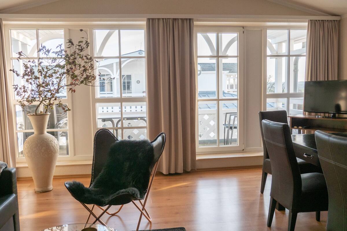 Ein modernes Wohnzimmer mit großem Fenster, einem schwarzen Ledersessel, einer weißen Vase mit Blumen und einem Flachbildfernseher auf einem Holzständer.