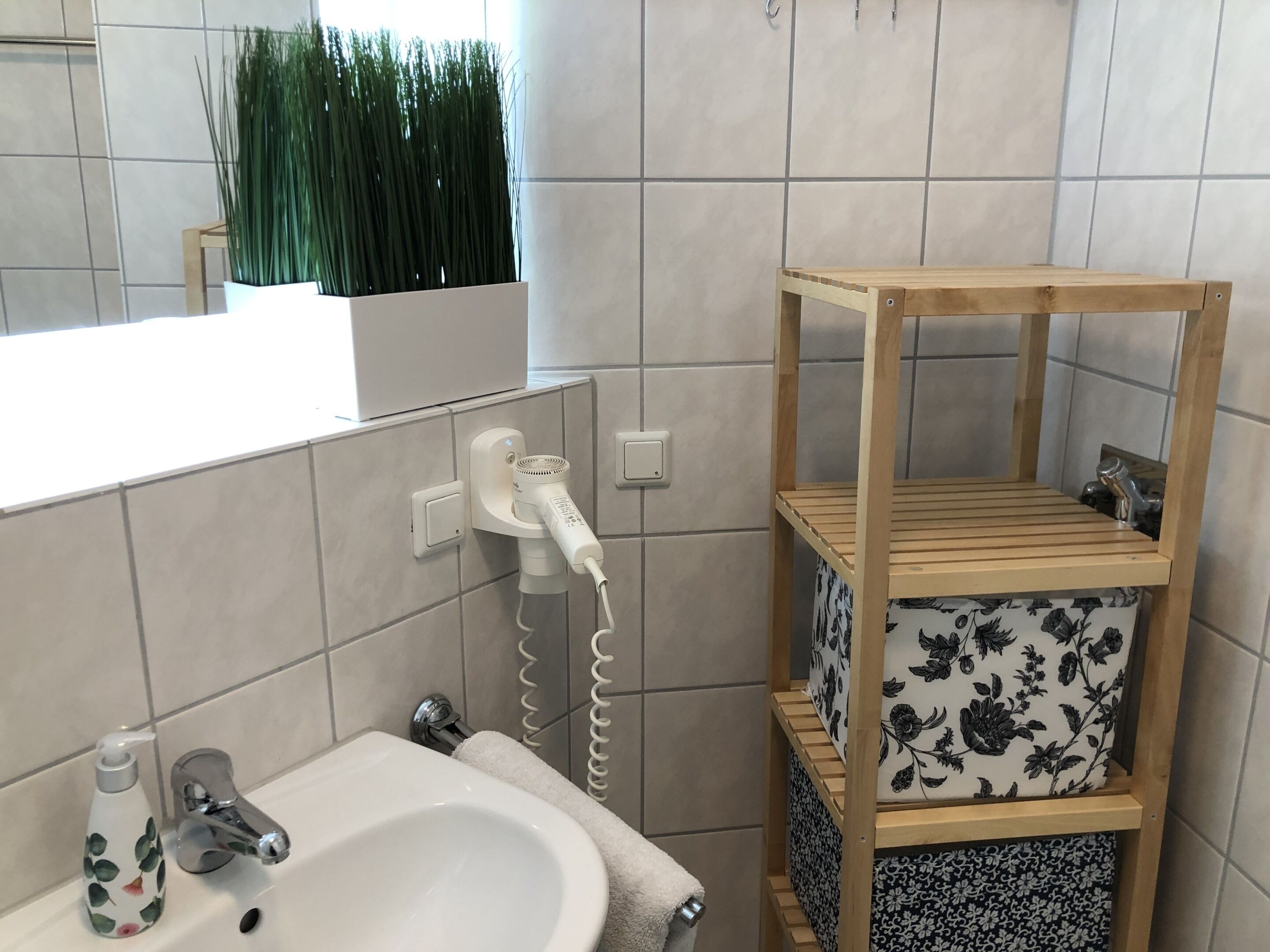 Un baño con un lavabo blanco, un espejo, una toalla, un secador de pelo y una estantería de madera.