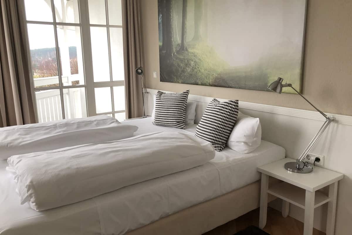 Ein ordentlich bezogenes Bett mit weißen Leinen und zwei gemusterten Kissen, ein Nachttisch mit einer Lampe und ein großes abstraktes Gemälde an der Wand.