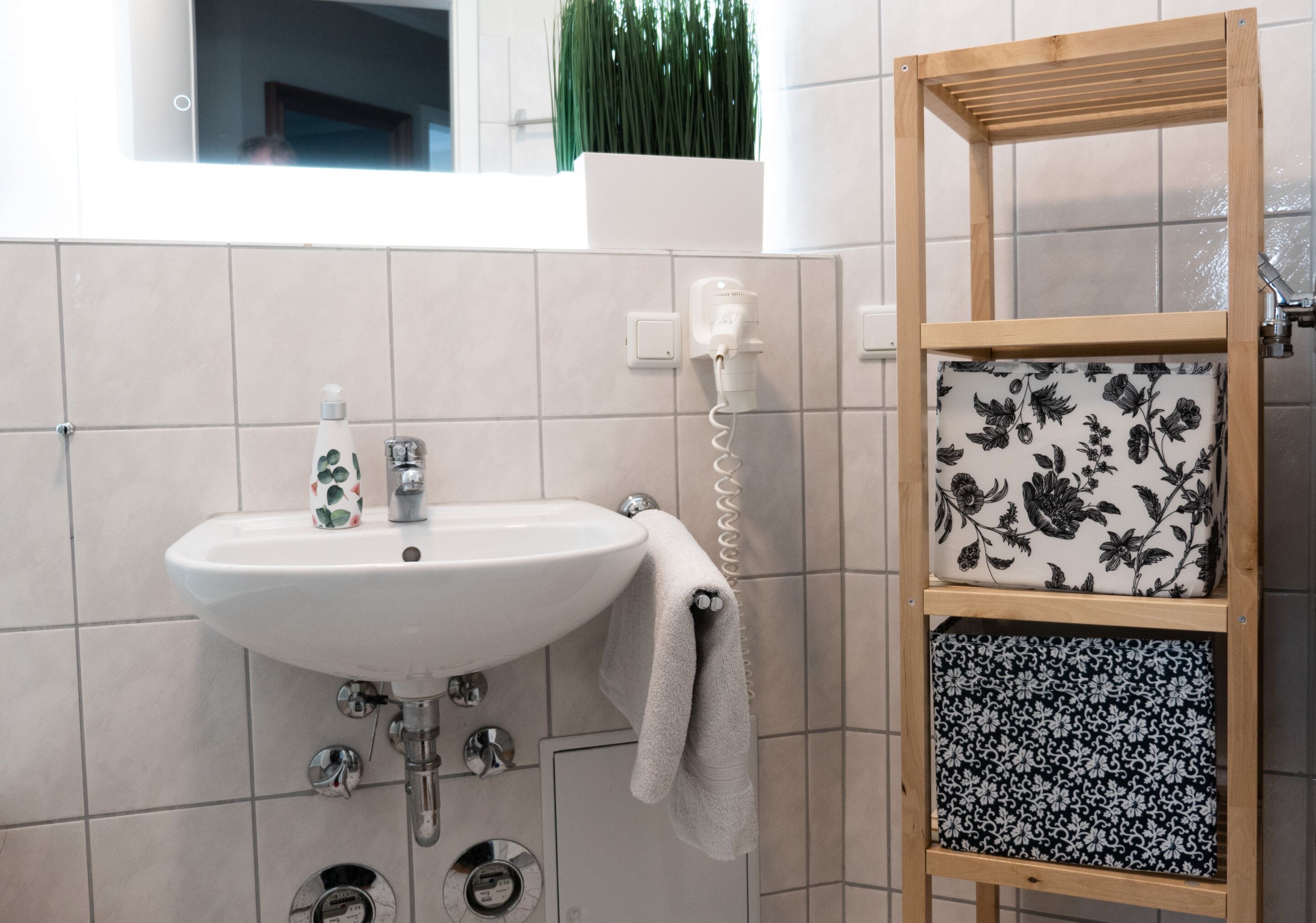 Een badkamer met een wit wastafel, een handdoek aan de muur, een houten plank met gestreken stof en een groene plant op de achtergrond.