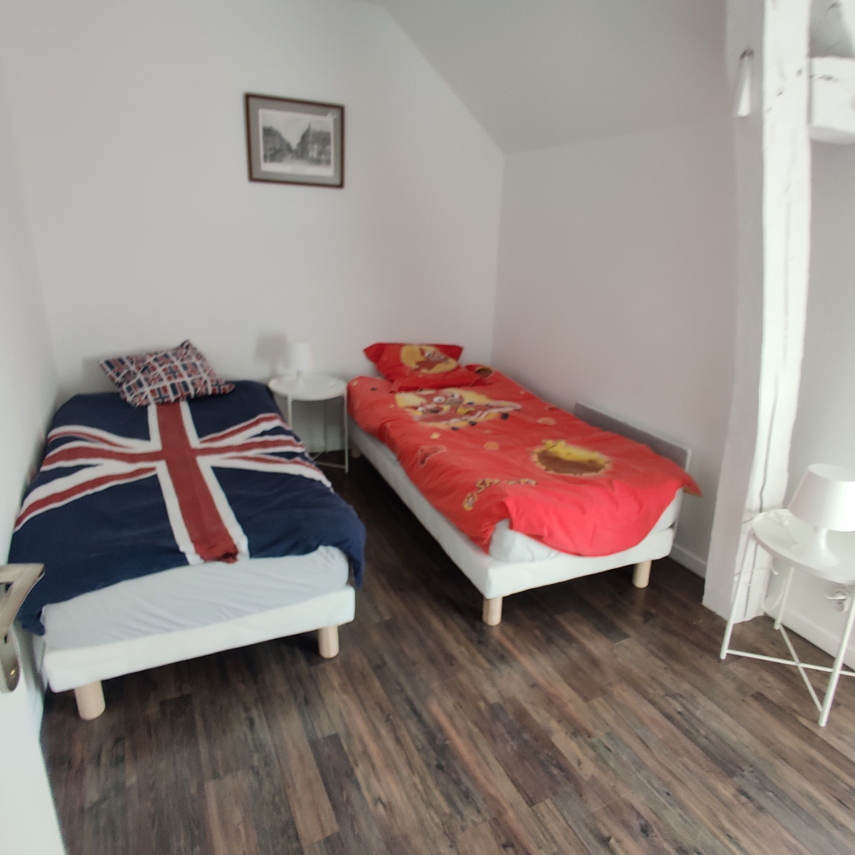 Un dormitorio con dos camas individuales, una mesita de noche pequeña, una lámpara blanca y un cuadro en la pared.