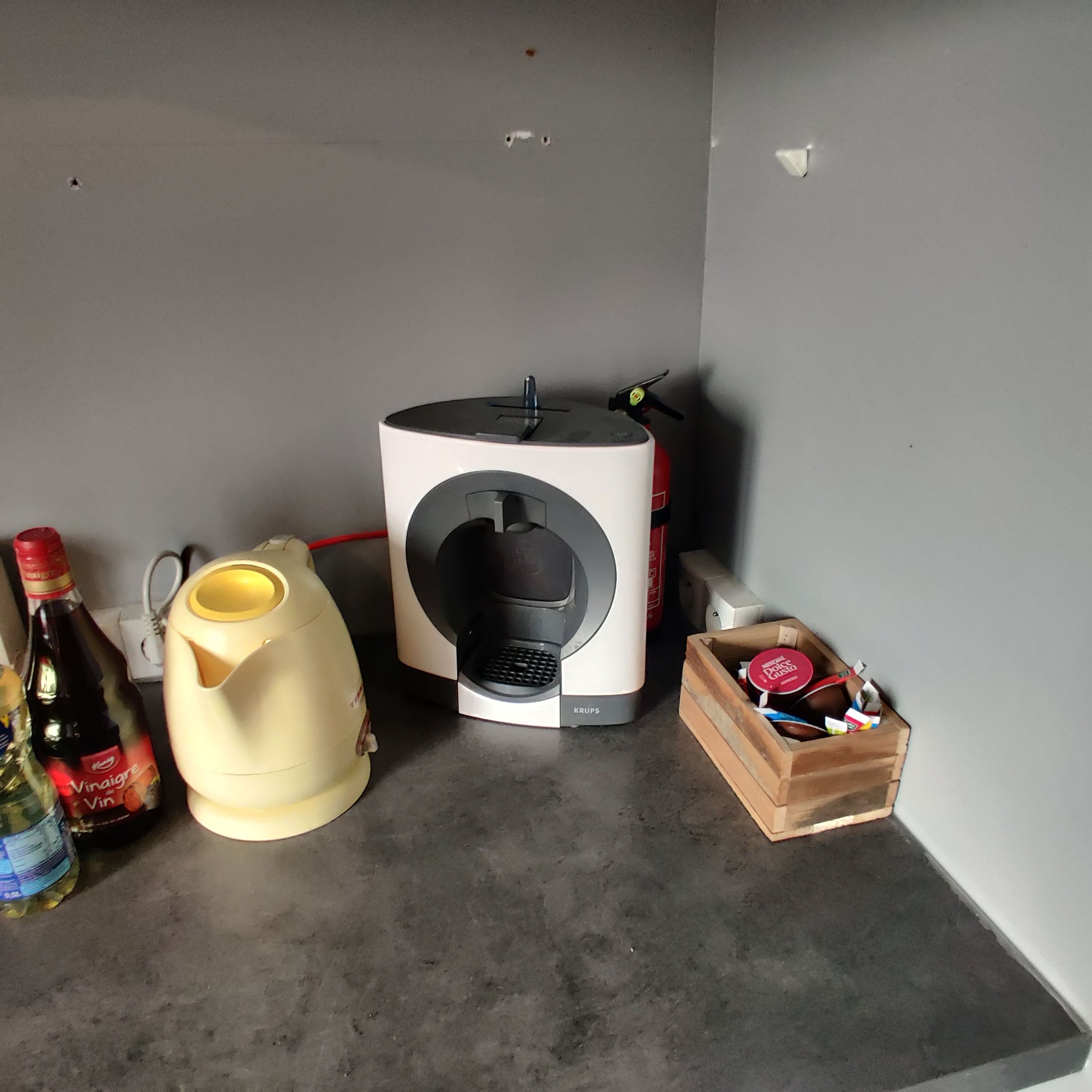 Een koffiezetapparaat, een fles Vinaigre de Vin, een gele kan en een houten doos met spullen op een aanrecht.
