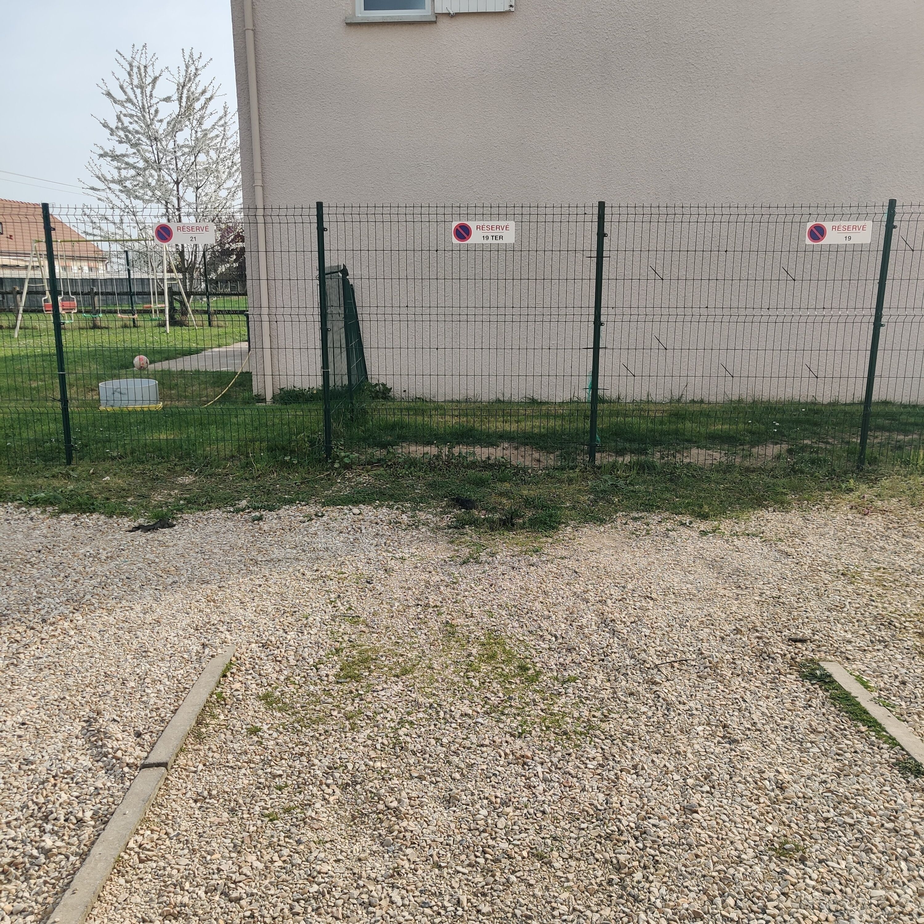 Un espace extérieur clos, avec un chemin de gravier, une clôture en métal et un bâtiment en arrière-plan.
