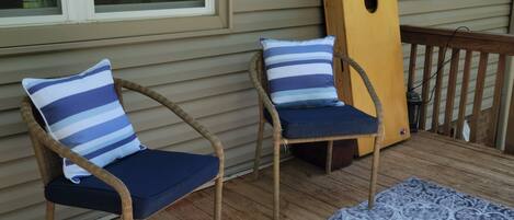 A wooden deck with two wicker chairs, each with a blue cushion and a striped pillow. A patterned rug is laid out on the deck.