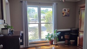 Ein Zimmer mit Deckenventilator, einem großen Fenster mit Vorhängen, Holzboden, einer Topfpflanze und einem Schrank.