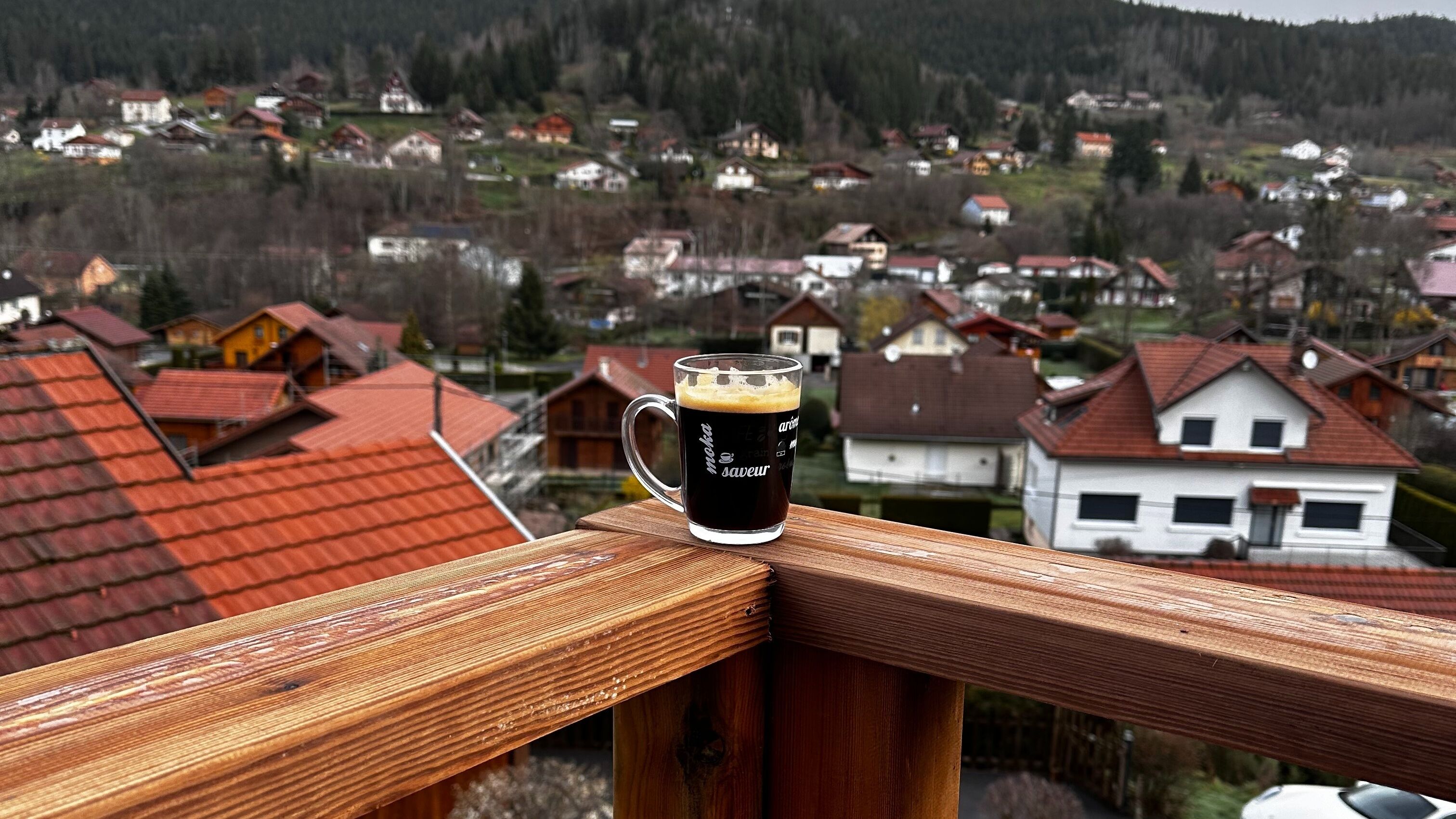 A glass of dark beer on a wooden railing overlooking a village with red-roofed houses.