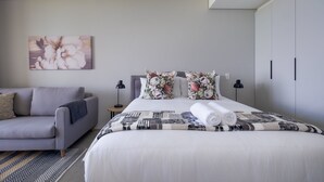 A modern hotel room with a large bed, a grey sofa, floral pillows, and a wall-mounted artwork. - Cosy Studio - Umhlanga Ridge - Pool - Aircon - Gym (Cape Town)