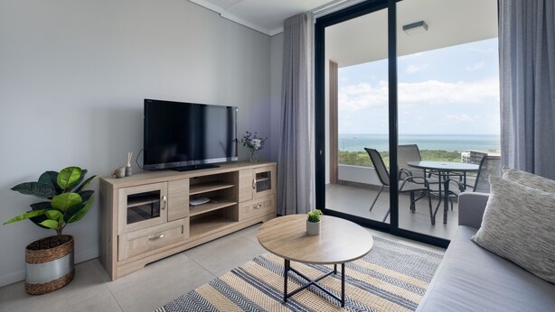 A modern living room with a TV on a wooden cabinet, a round coffee table, a sofa, and a balcony with a view of the sea. - Cosy Studio - Umhlanga Ridge - Pool - Aircon - Gym (Cape Town)