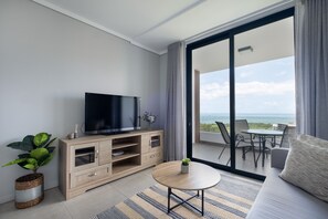 A modern living room with a TV on a wooden cabinet, a round coffee table, a sofa, and a balcony with a view of the sea. - Cosy Studio - Umhlanga Ridge - Pool - Aircon - Gym (Cape Town)