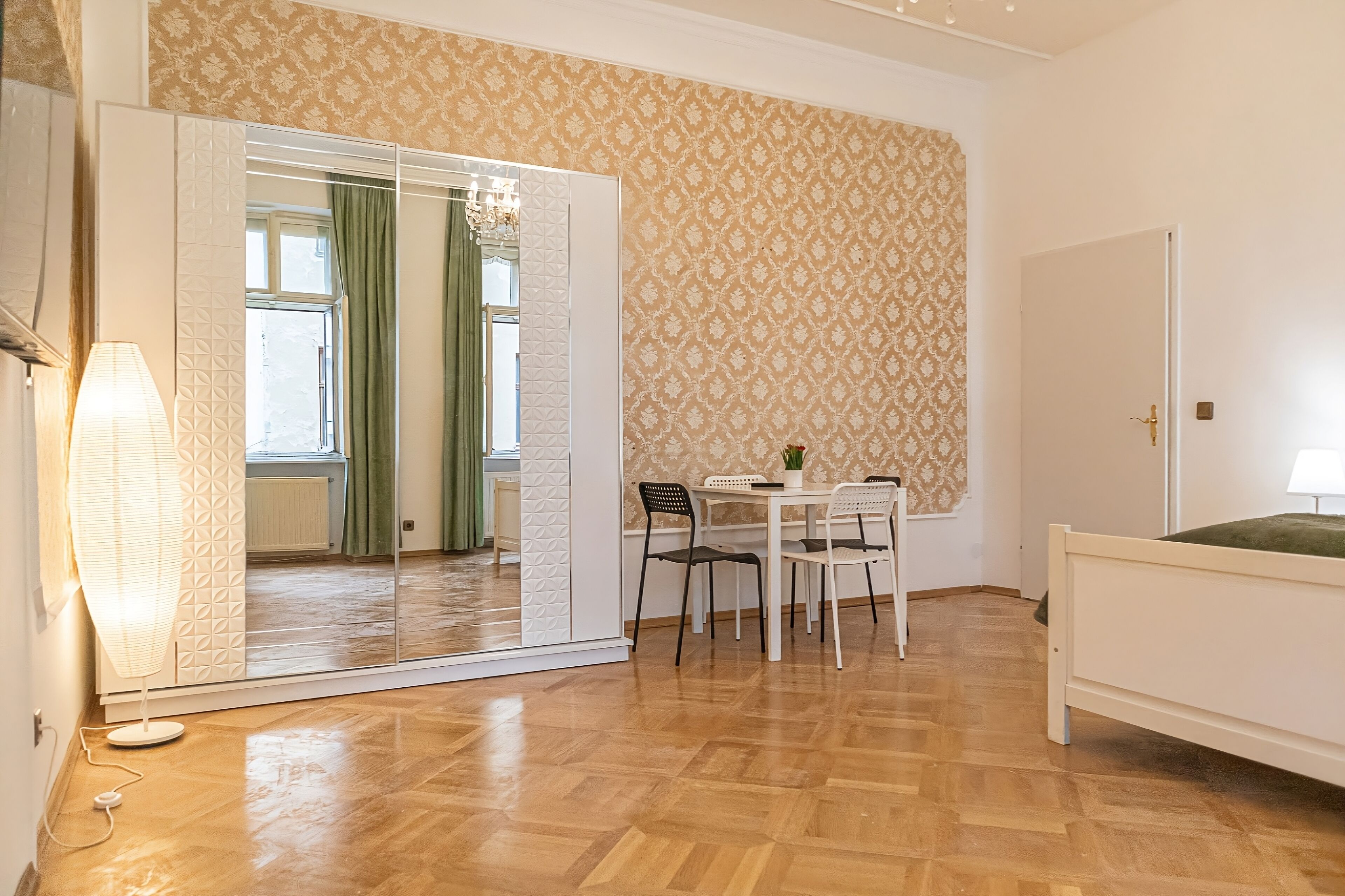 Ein Zimmer mit Holzboden, einem Bett, einem Tisch mit Stühlen, einem Spiegel und einem Wandpapier mit Muster.