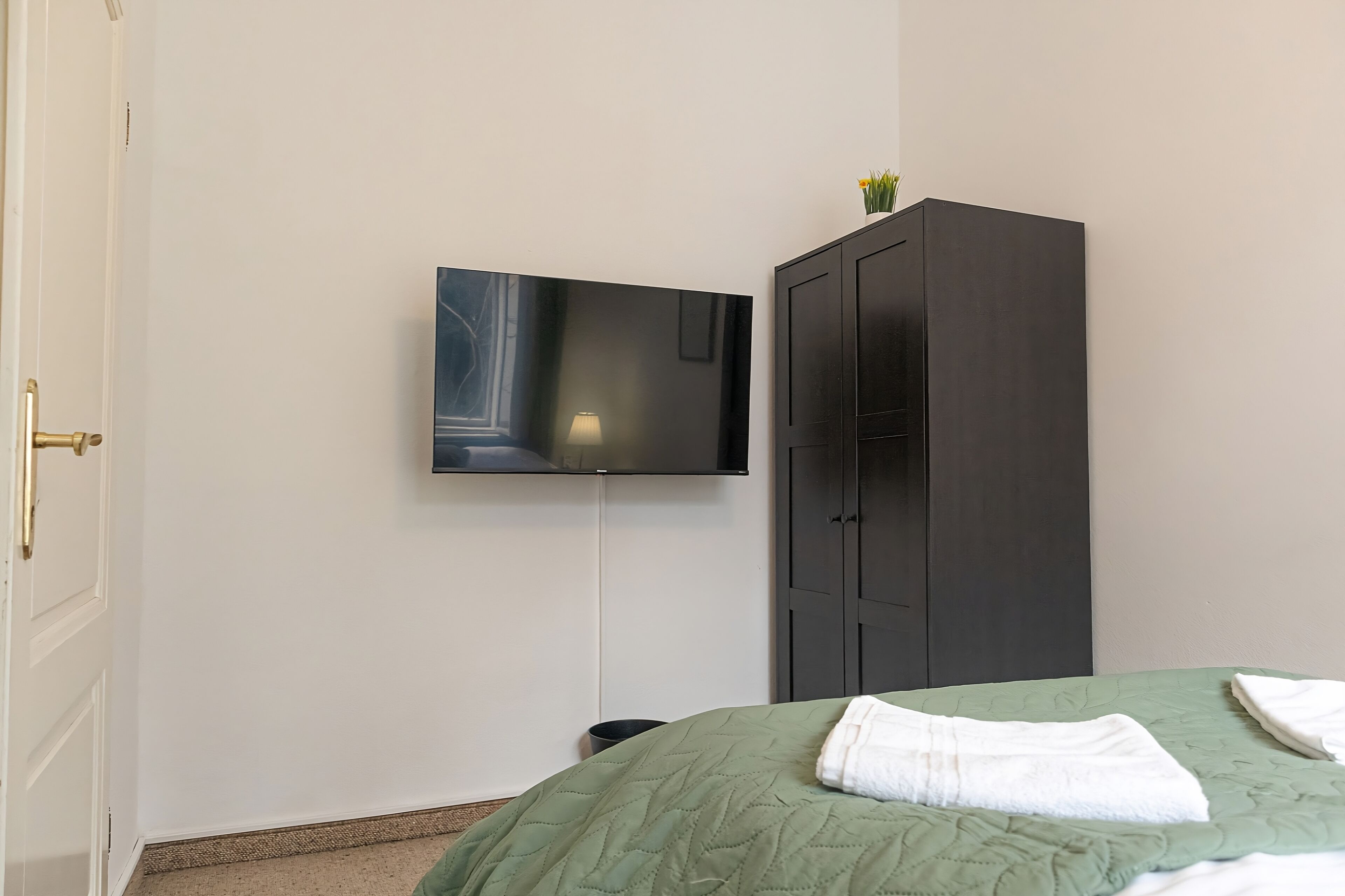 A room with a bed, a dark wardrobe, a mounted TV, and a small table with a lamp.