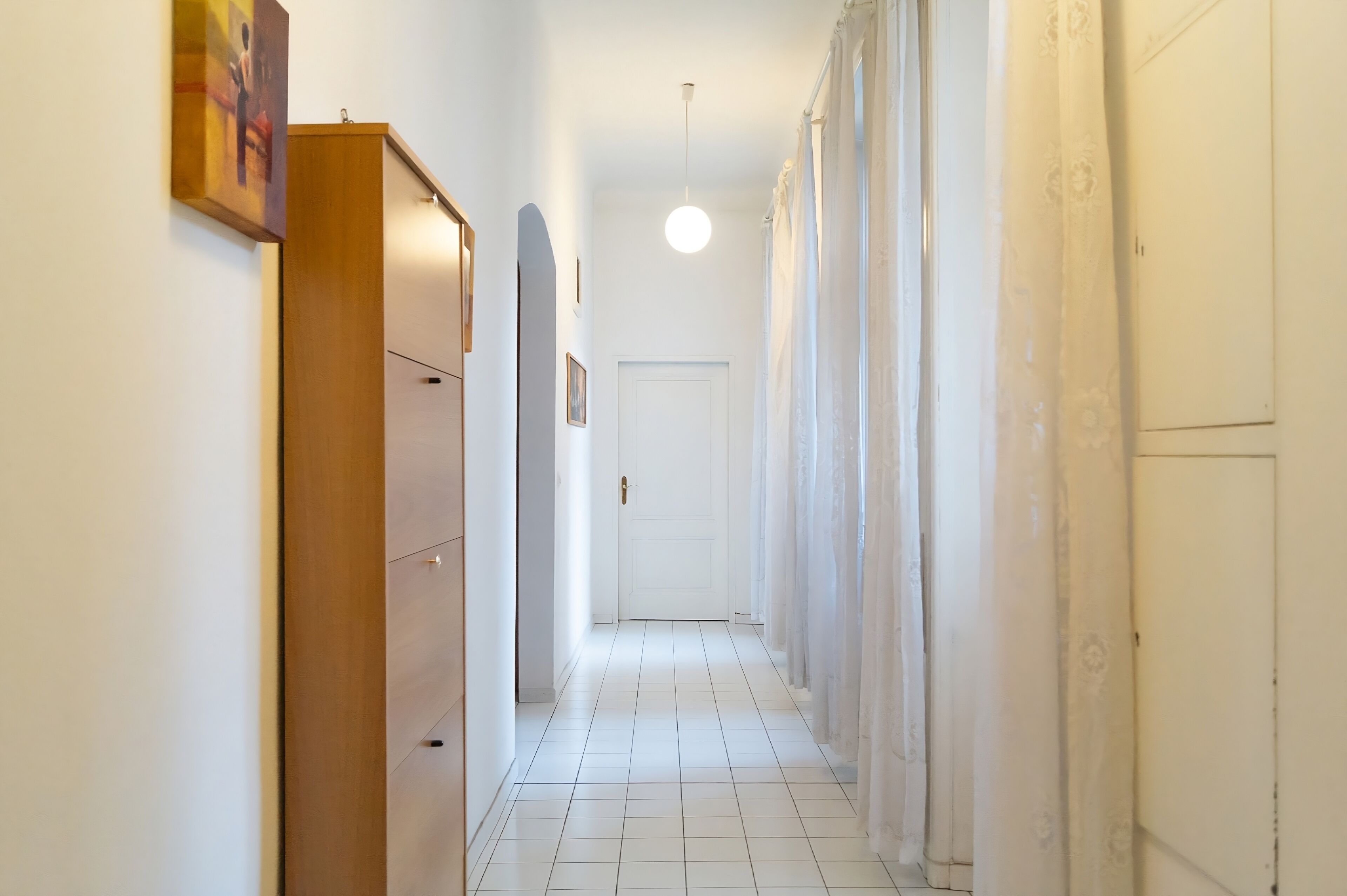 A narrow hallway with white tiled flooring, a wooden wardrobe on the left, a white door at the end, and a hanging light fixture.