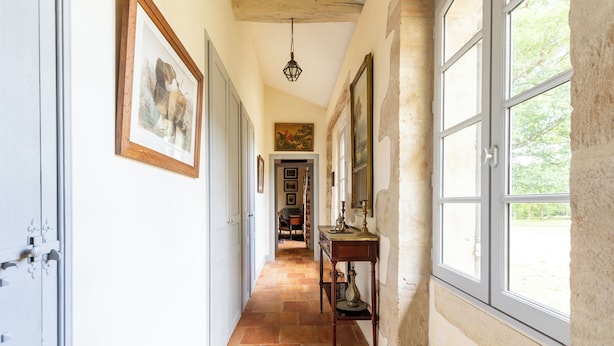 Un couloir doté d’un sol carrelé, de tableaux encadrés accrochés aux murs, d’un bureau avec une lampe et de grandes fenêtres.