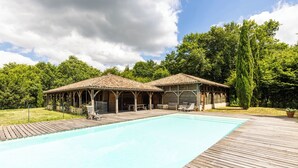 Een houten terras met een zwembad, twee overdekte houten constructies met zitplaatsen, en een achtergrond van bomen en een bewolkte lucht.