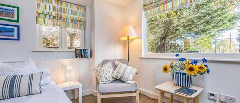 A bedroom with a bed, a chair, a bedside table with a lamp, a window with curtains, and a vase with flowers.
