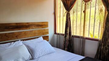 Una cama con ropa de cama blanca y una manta oscura, cabecero de madera y una ventana con cortinas.