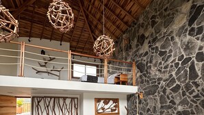 Un dormitorio con pared de piedra, techo de madera, cama con ropa de cama gris, sofá gris y una lámpara colgante.