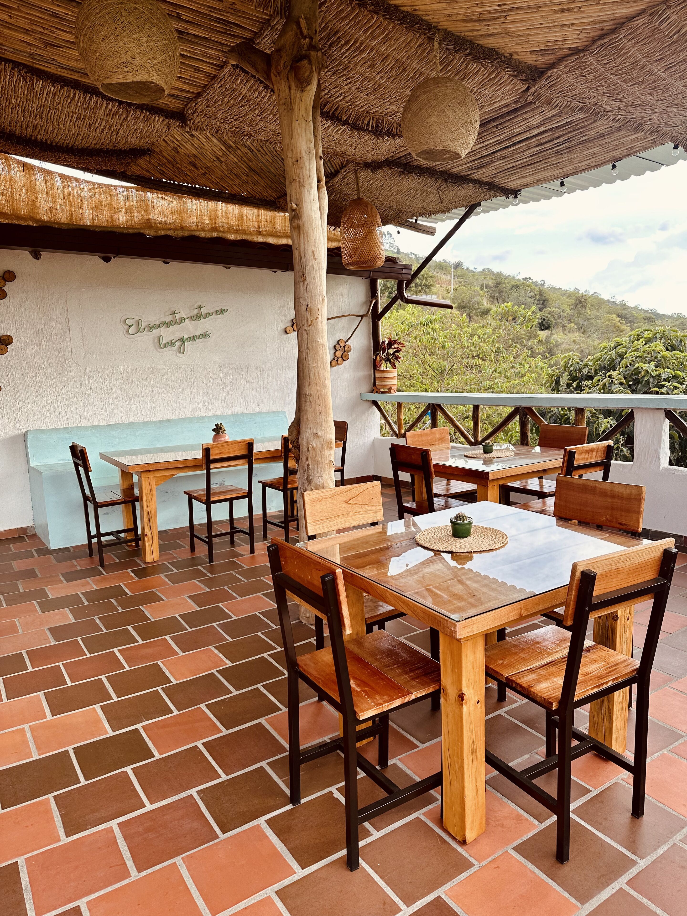 An outdoor dining area with wooden tables and chairs, a thatched roof, and a view of greenery in the background.