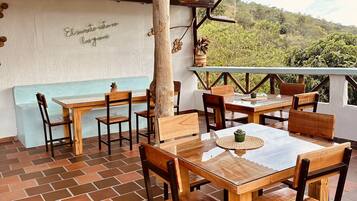 An outdoor dining area with wooden tables and chairs, a thatched roof, and a view of greenery in the background.