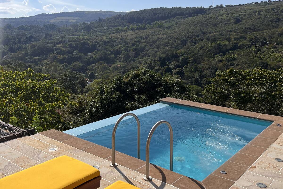 Ein Pool im Infinity-Stil mit Metallleitern, zwei gelben Liegestühlen und Blick auf einen bewaldeten Hang.