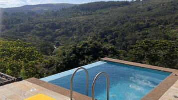 Una piscina infinita con escaleras de metal, dos sillas de descanso amarillas y vistas a una ladera boscosa.
