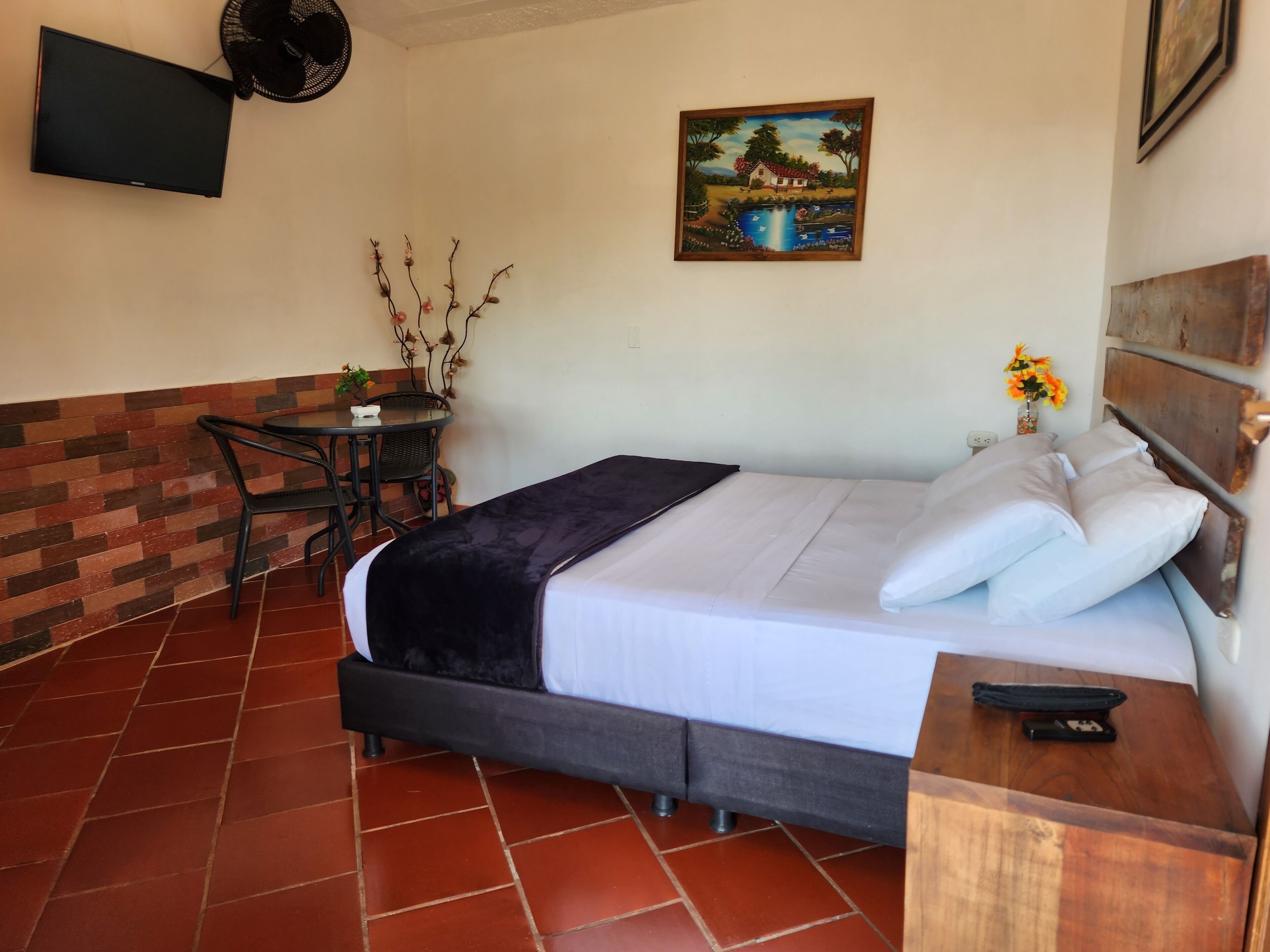 Una camera d'albergo con un letto, un tavolino, una sedia, un quadro appeso al muro e una TV a parete.