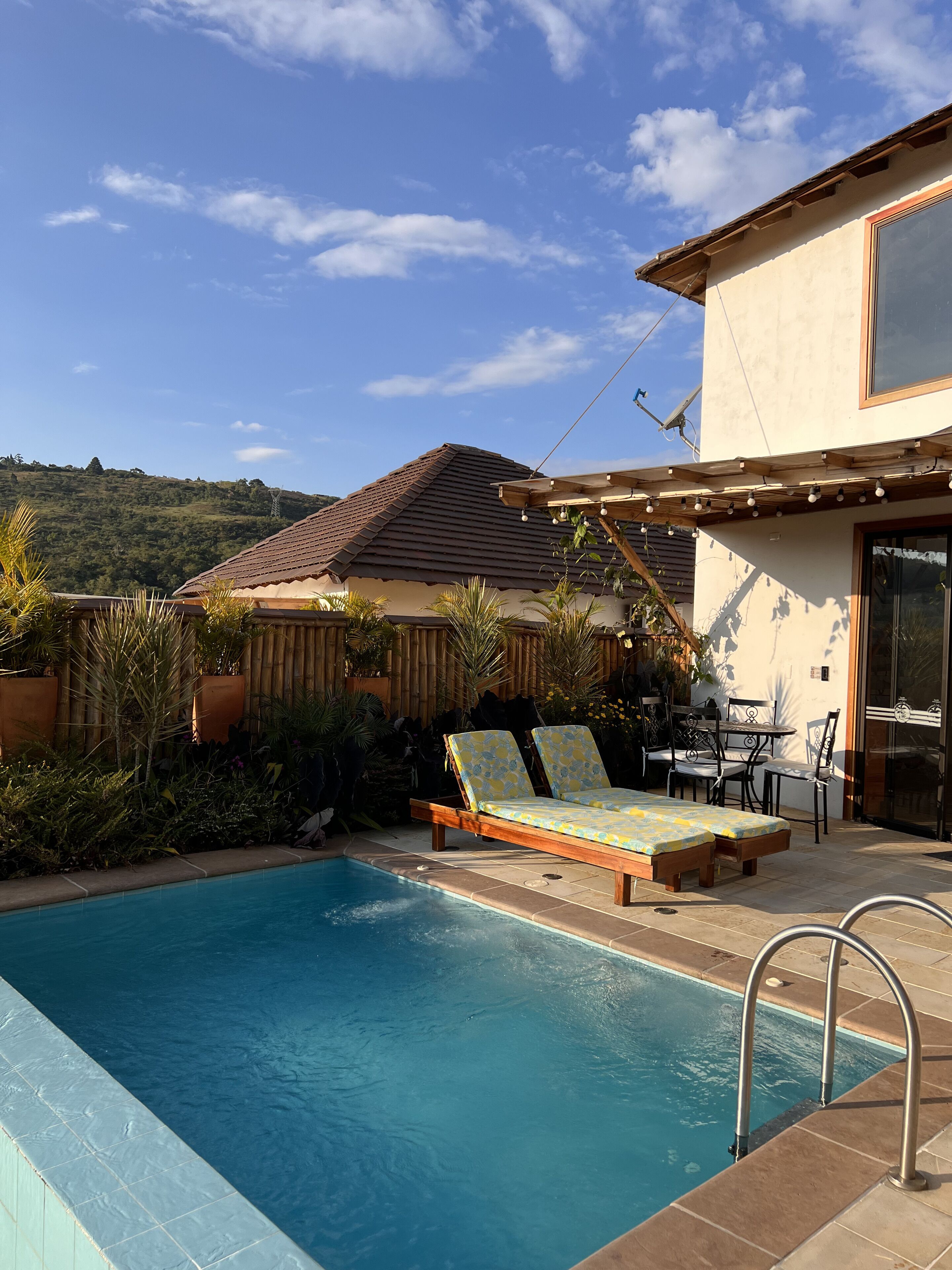 Une piscine entourée de plantes, avec des chaises longues et une table, et une colline en arrière-plan.