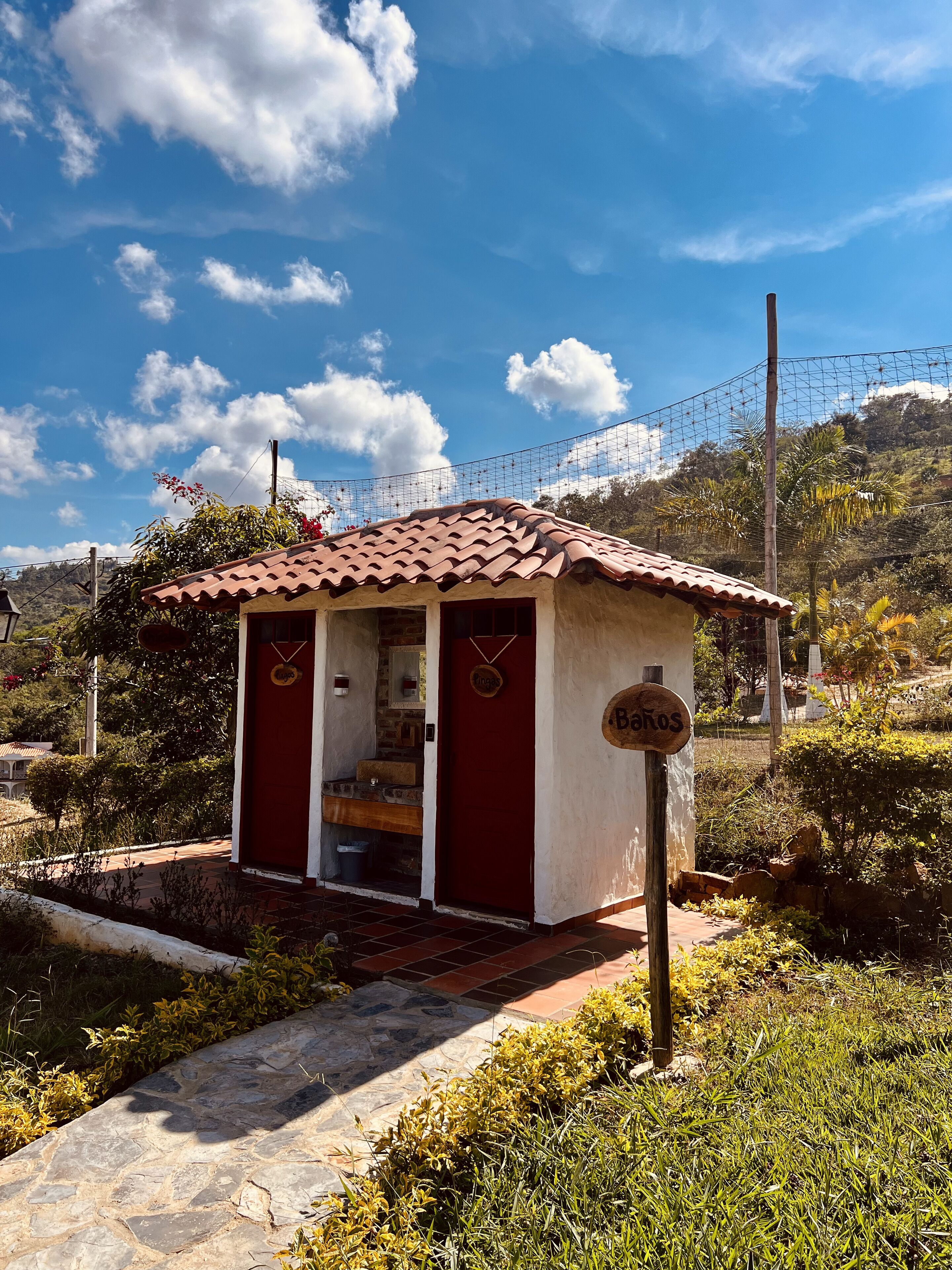 A small, rustic building with a tiled roof and a red door, surrounded by greenery and a clear sky.