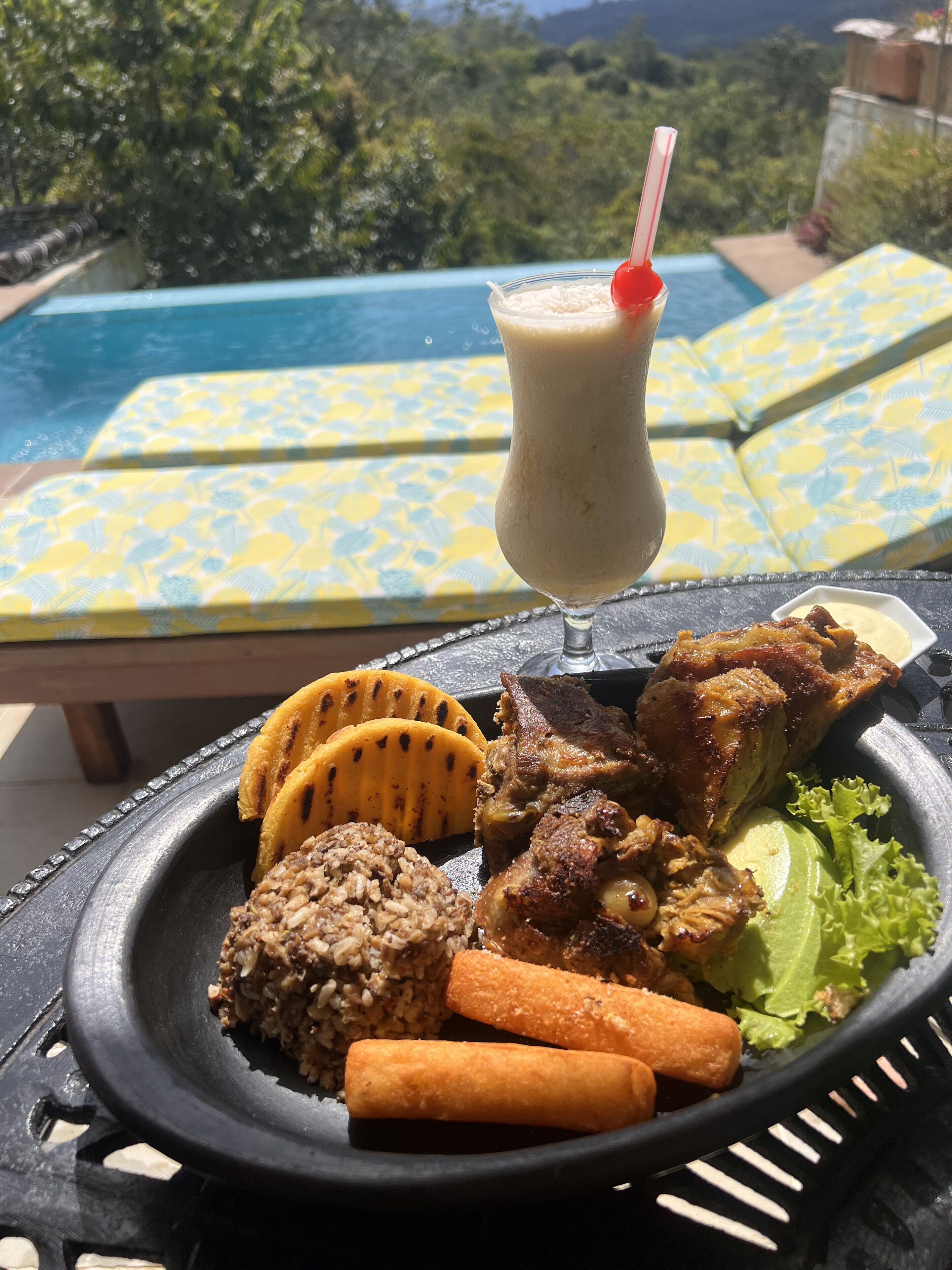 Una mesa de comedor con una comida que incluye carne a la parrilla, arroz y verduras, junto a una bebida con pajita. La mesa está al aire libre, con una piscina y sillas de descanso al fondo.