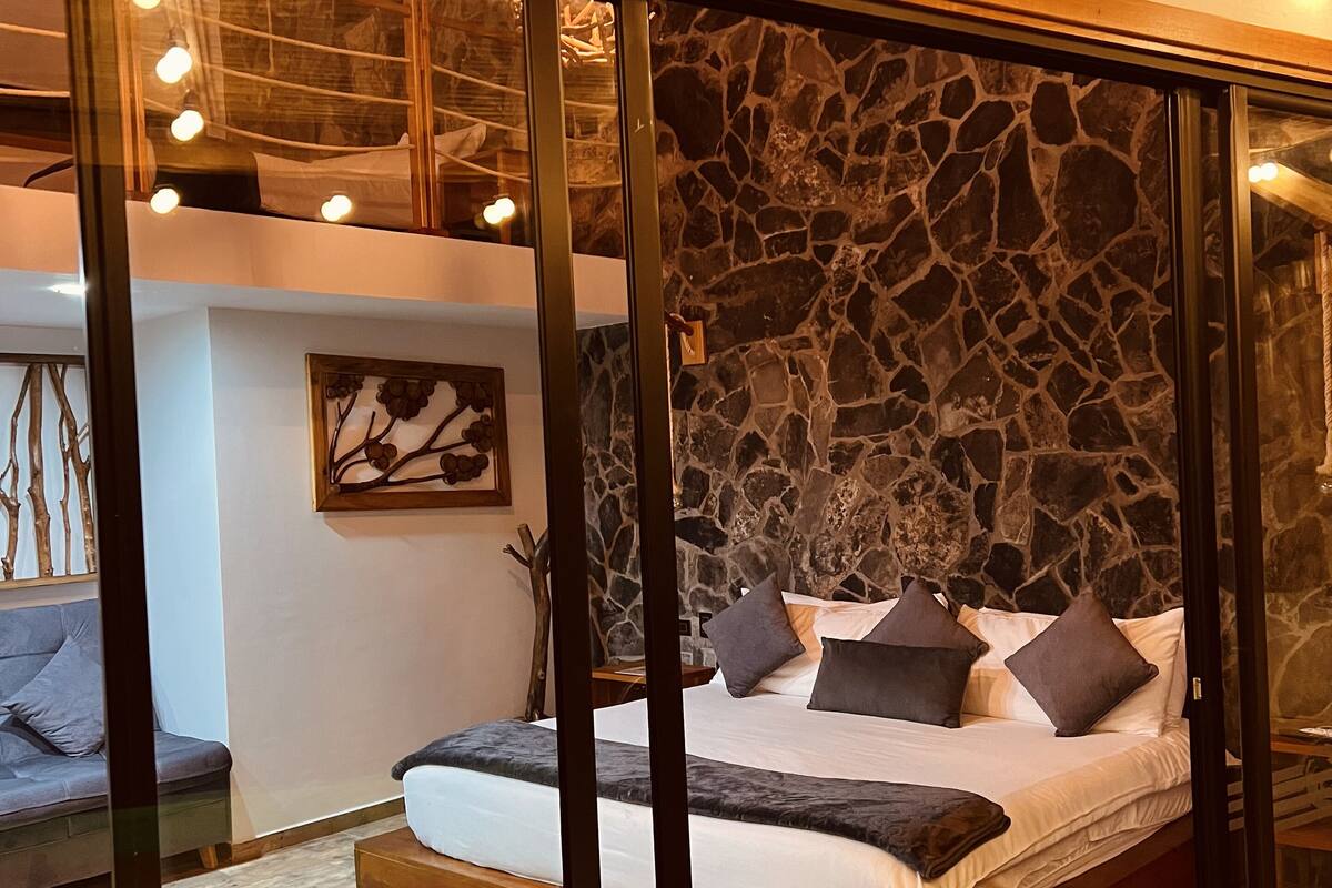Ein Schlafzimmer mit Steinsandsteinwand, einem Bett mit Kissen, Holzboden und einer Decke mit freiliegenden Balken und Lampen.