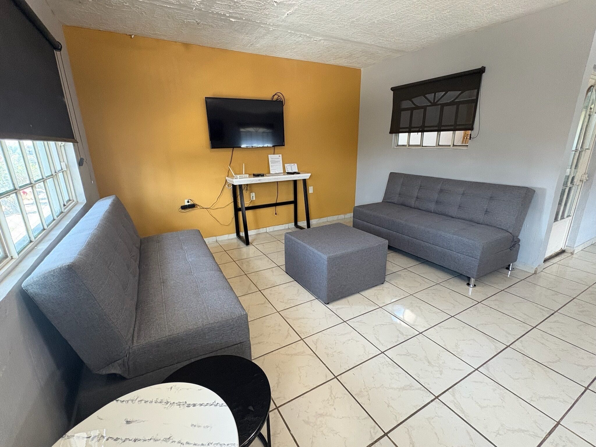 A living room with a grey sofa, a matching ottoman, a flat-screen TV mounted on the wall, a small table, and a window with bars.