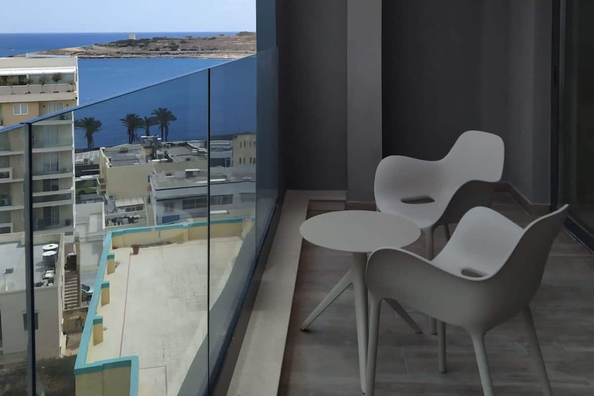 A balcony with two white chairs and a small table, overlooking a coastal area with buildings and a clear sky.
