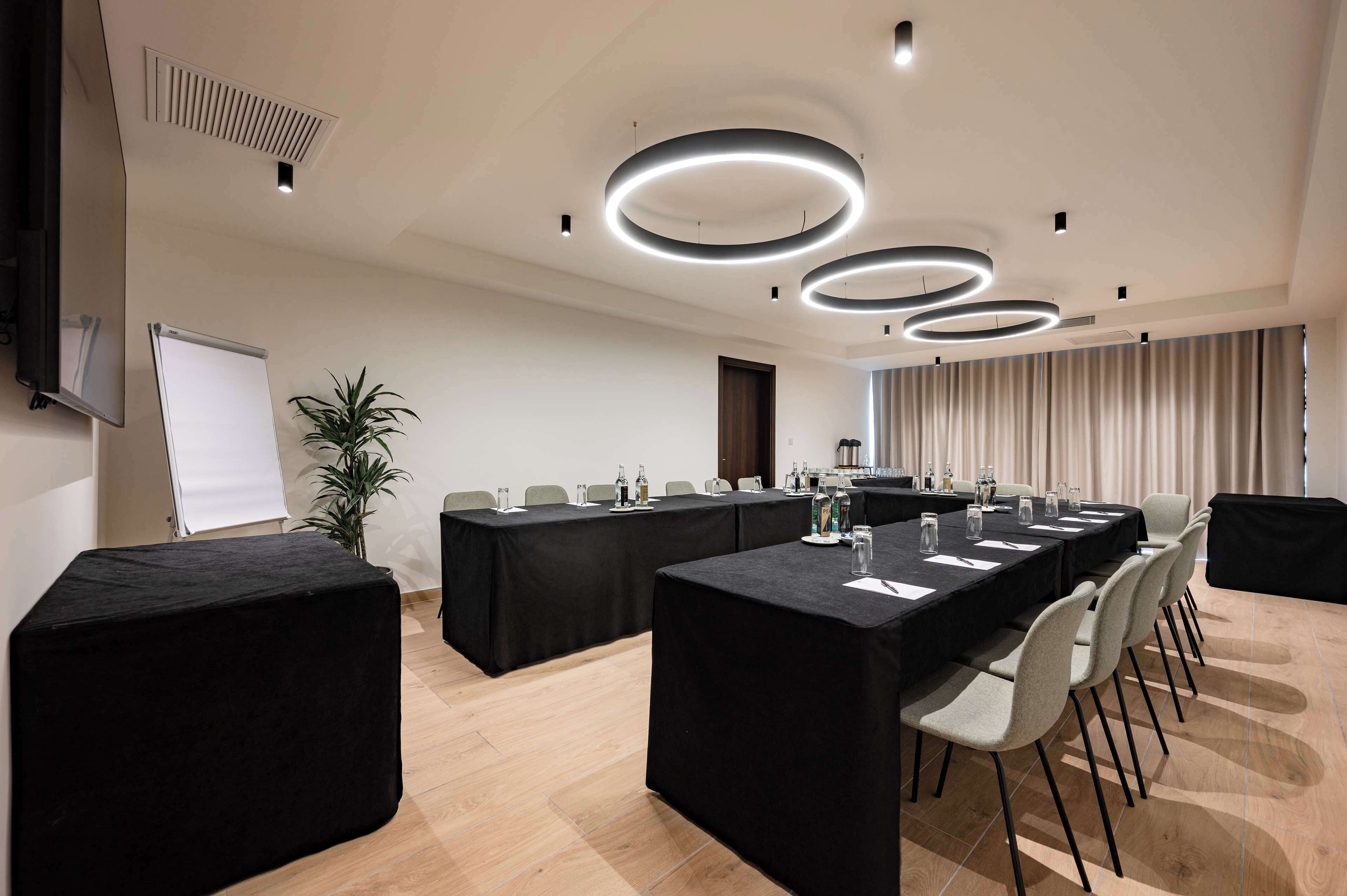 Un salón de conferencias con una larga mesa, manteles negros y modernas lámparas circulares en el techo.