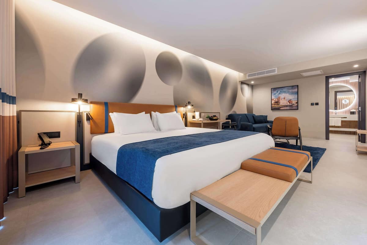 A modern hotel room with a large bed, bedside tables, a bench, a desk, and a chair.