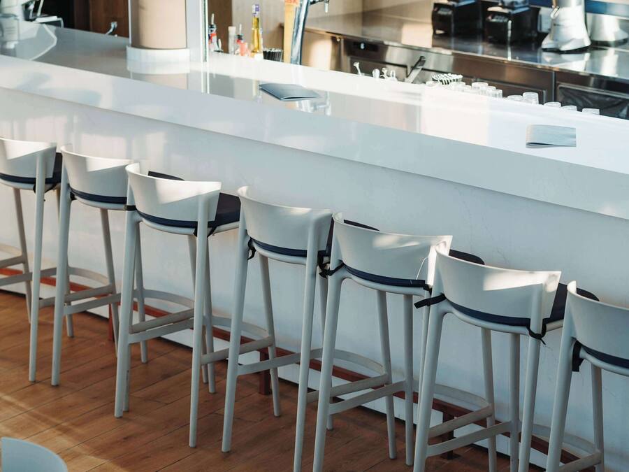 A modern bar area with white bar stools, a long counter, and a variety of bottles and glasses.