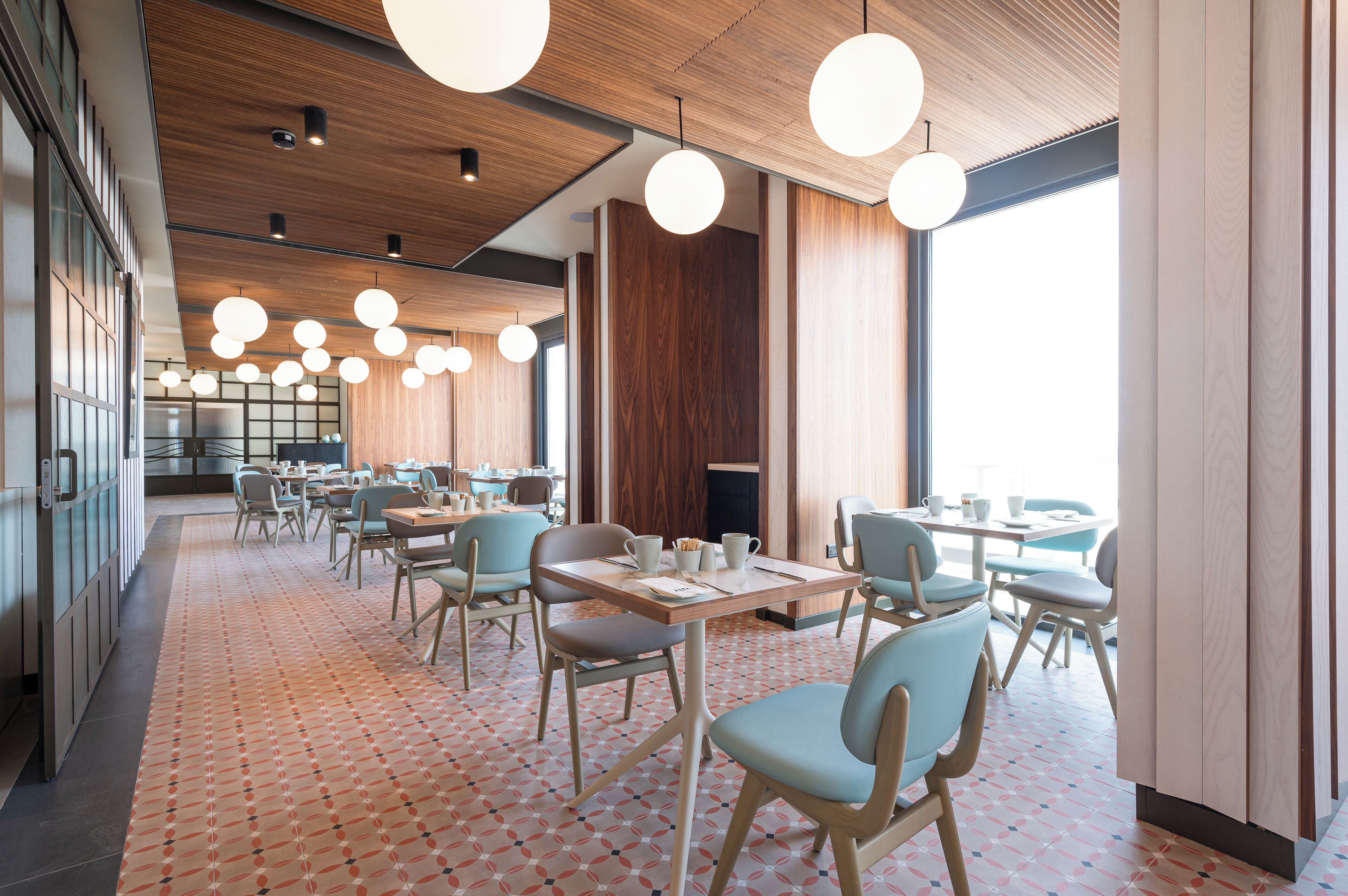 A modern restaurant with wooden ceiling panels, round pendant lights, and a patterned floor. Tables and chairs are arranged for dining.