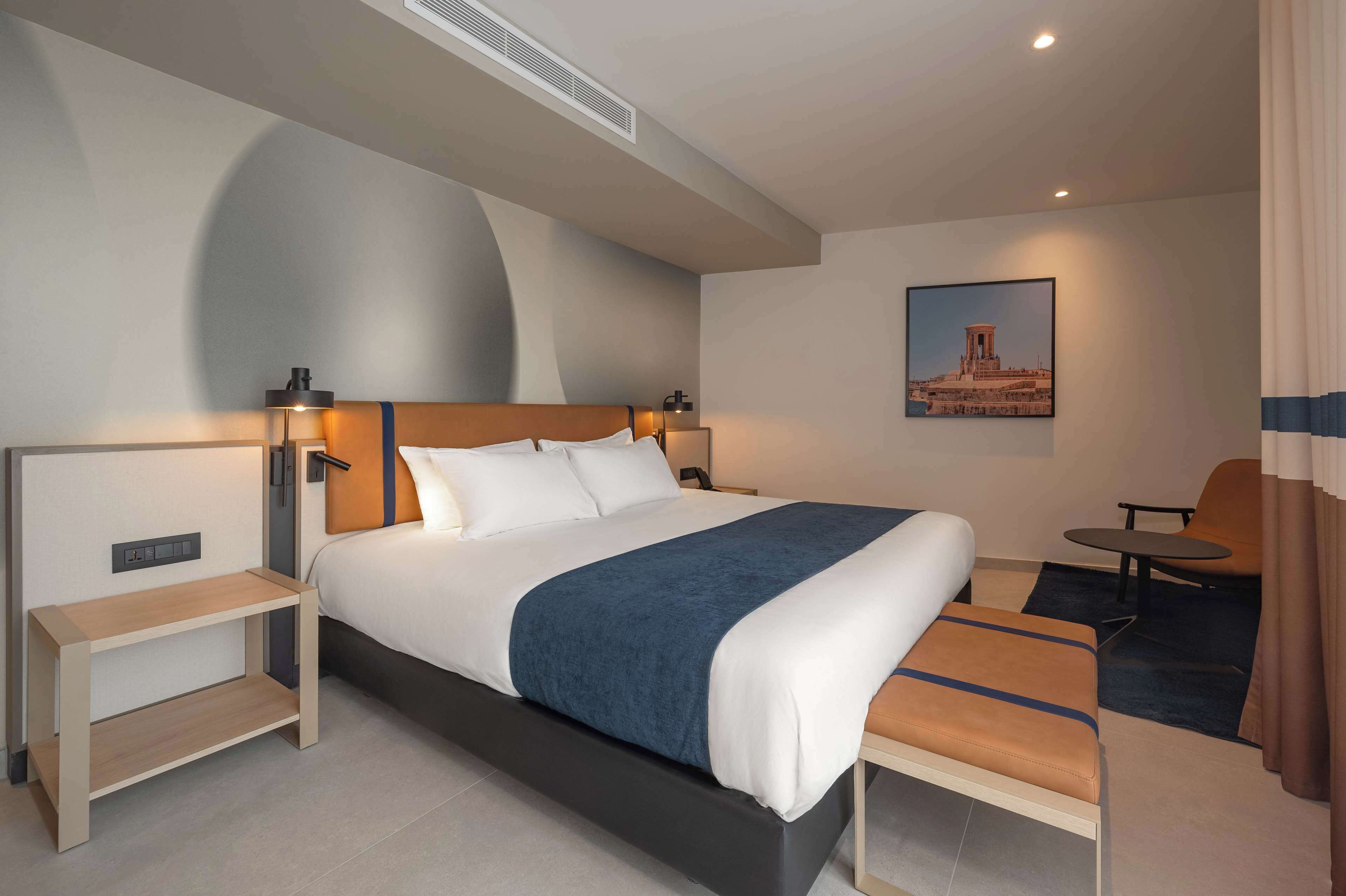 Una habitación de hotel moderna con una cama grande, mesitas de noche, un escritorio pequeño, una silla y una mesa redonda.