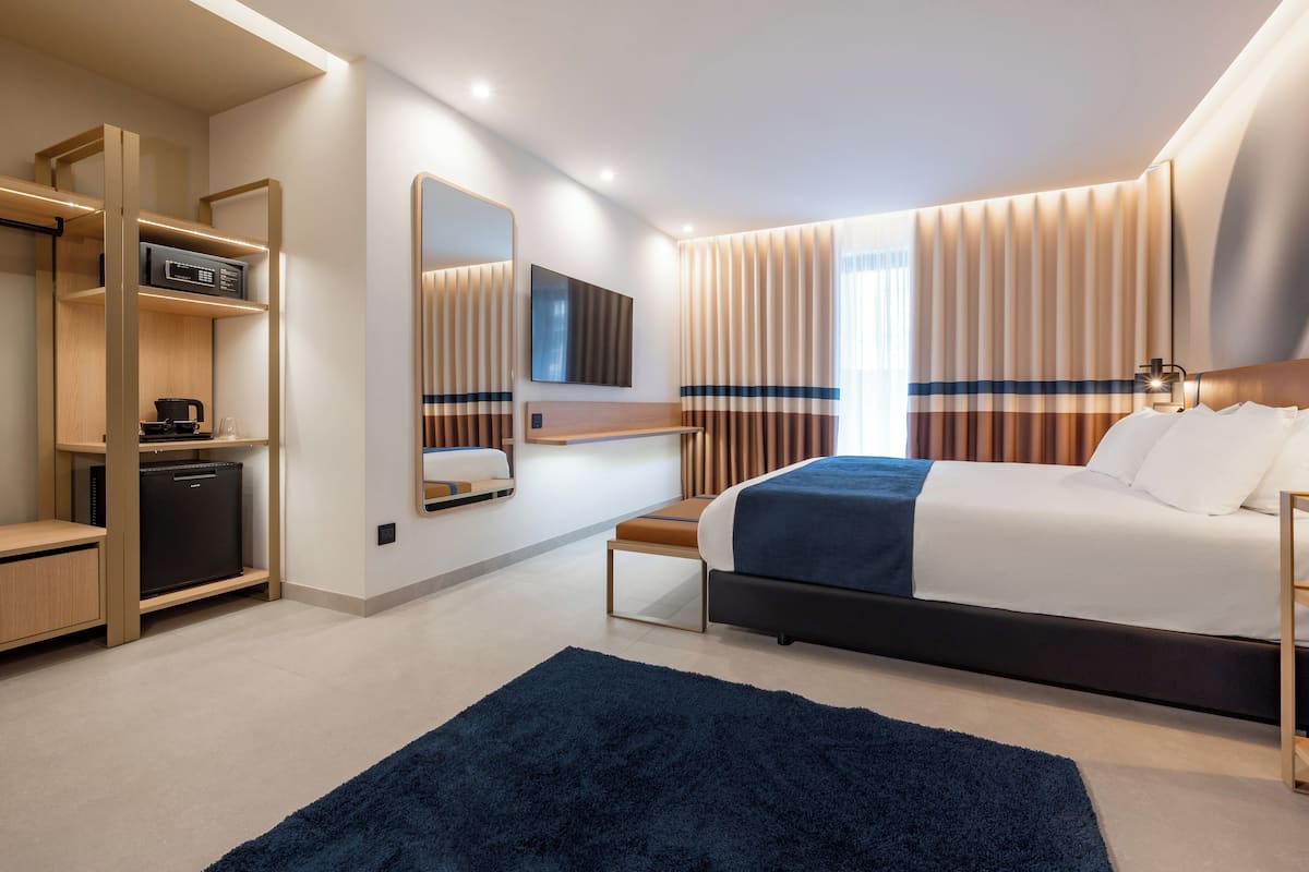 A modern hotel room with a large bed, a desk, a built-in TV, and a minibar.