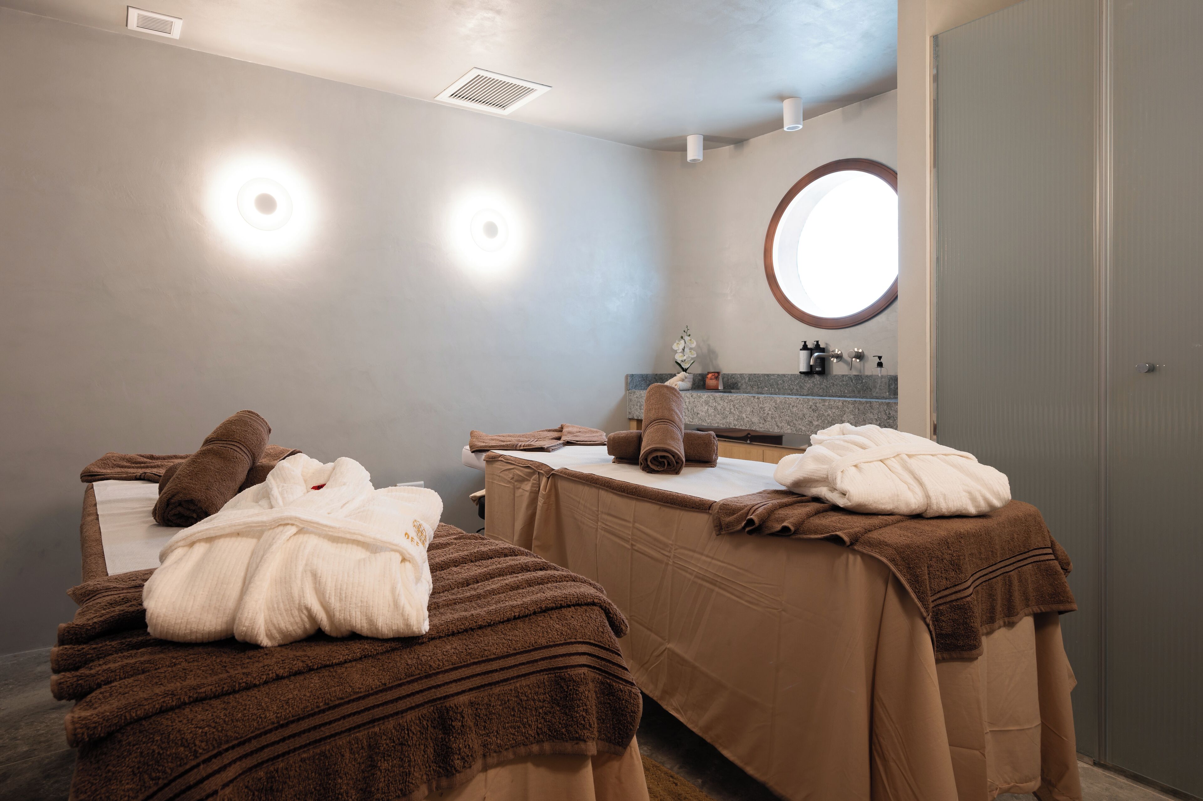 A spa room with two massage tables, towels, a mirror, and a sink.