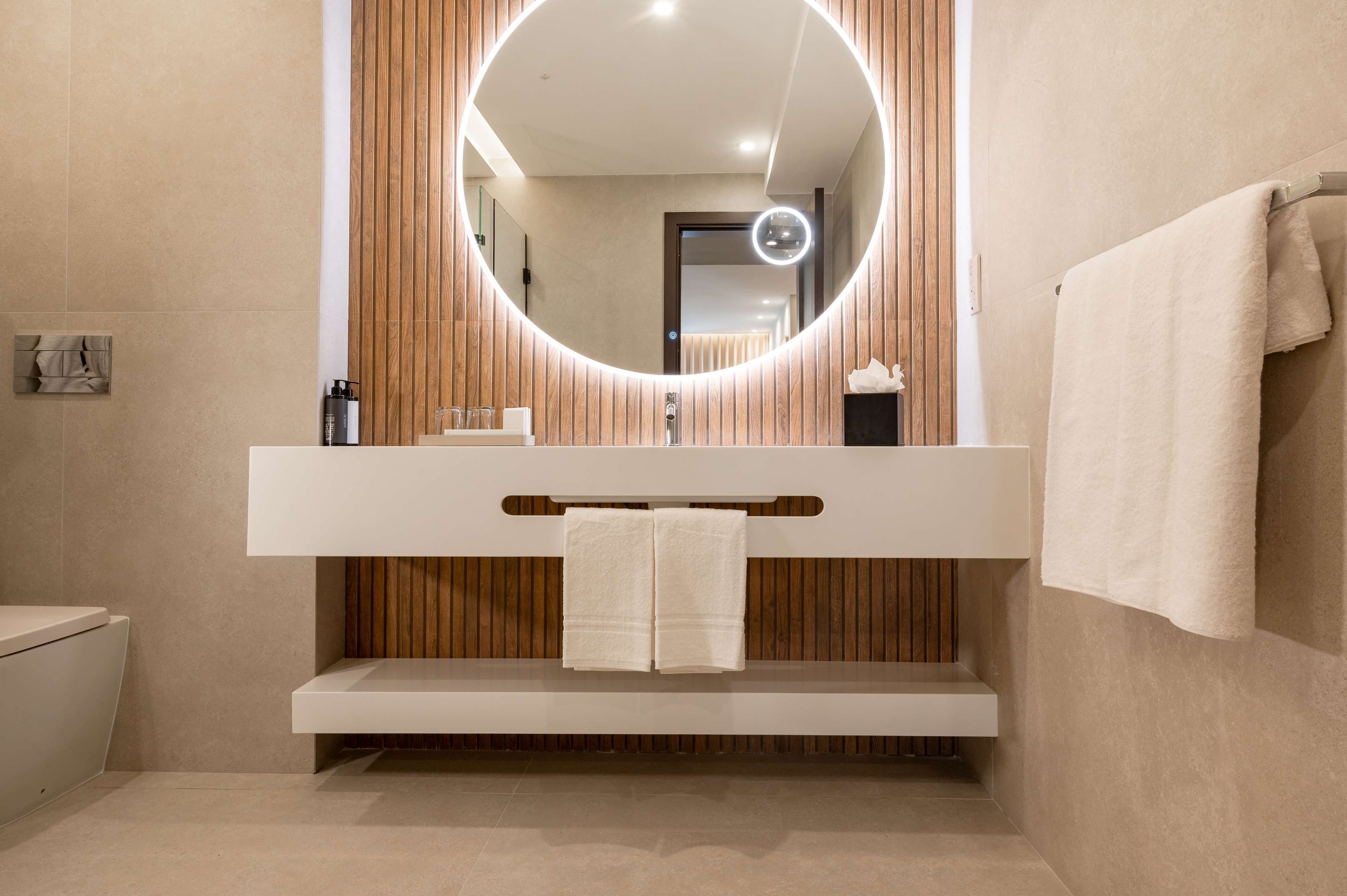 Une salle de bain moderne avec un mur recouvert de panneaux en bois, un grand miroir rond doté d’un éclairage intégré, un lavabo blanc et un porte-serviettes.