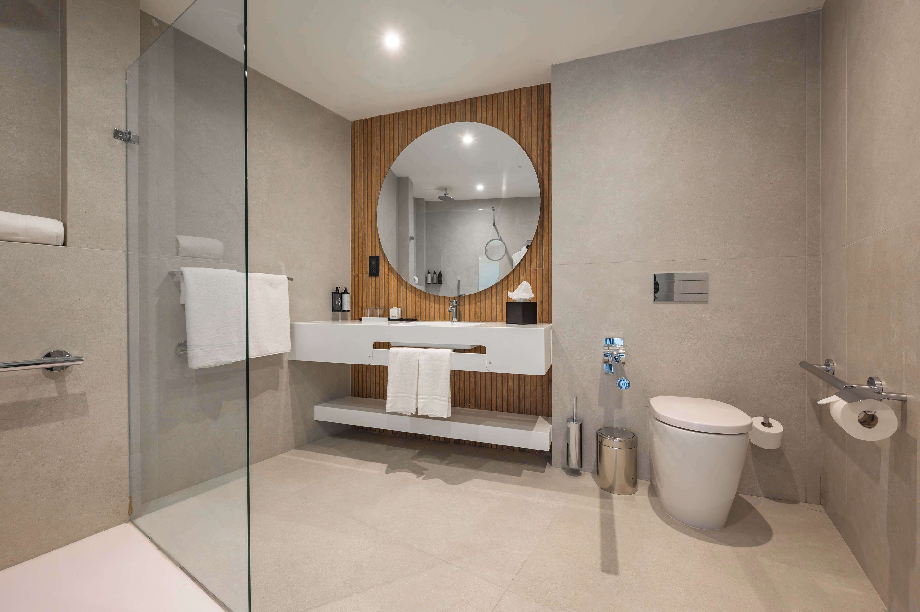 A modern bathroom with a glass shower enclosure, a wooden vanity, a round mirror, a toilet, and a bidet.