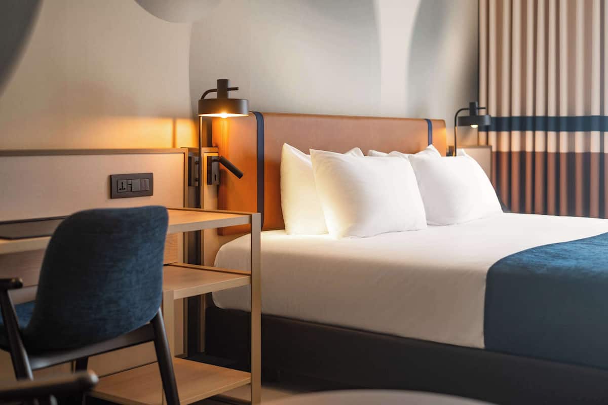 A hotel room with a bed, a desk, a chair, and a wall-mounted lamp.