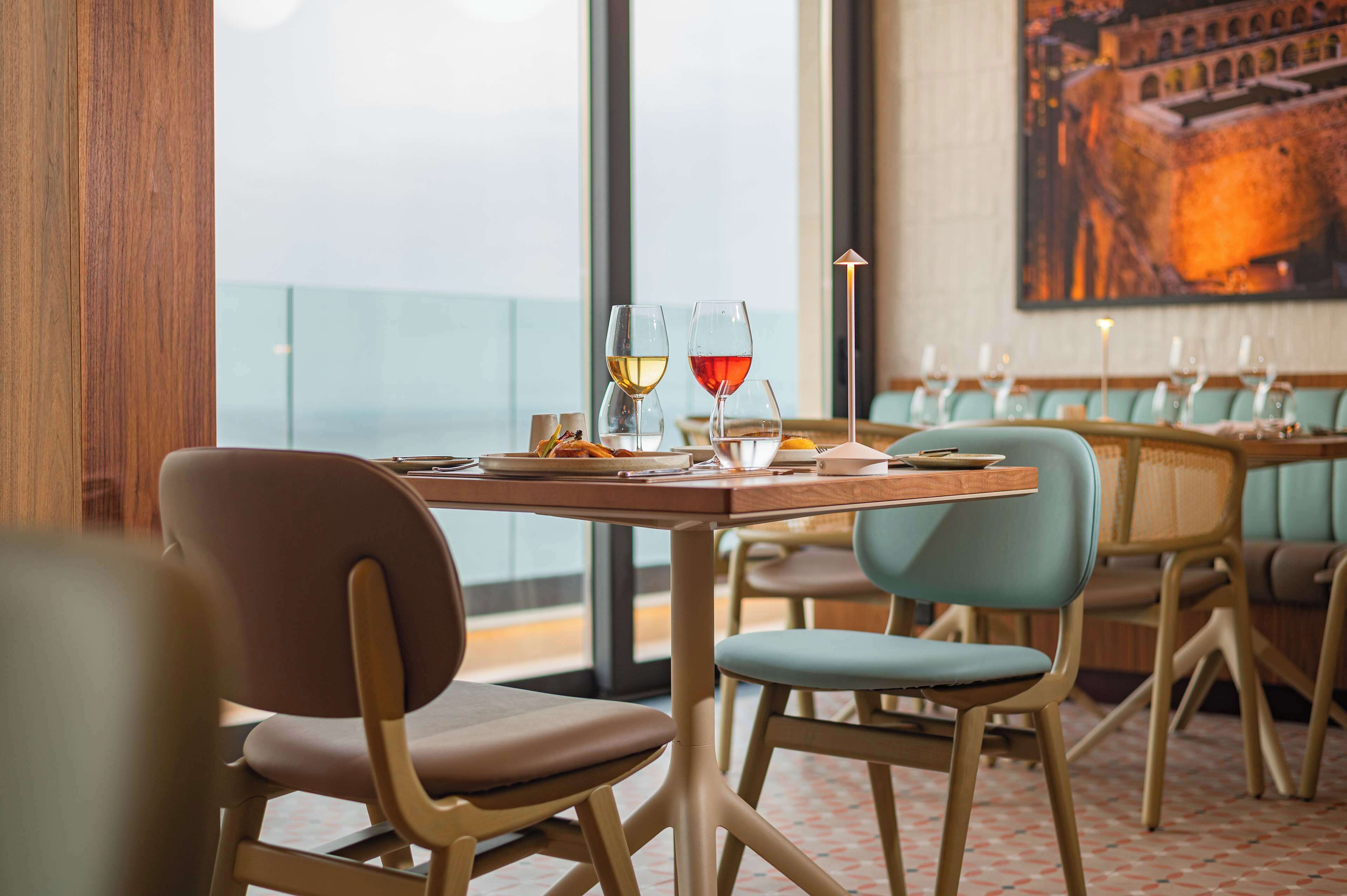 A restaurant with a view of the sea, wooden chairs, and tables with glasses of wine.