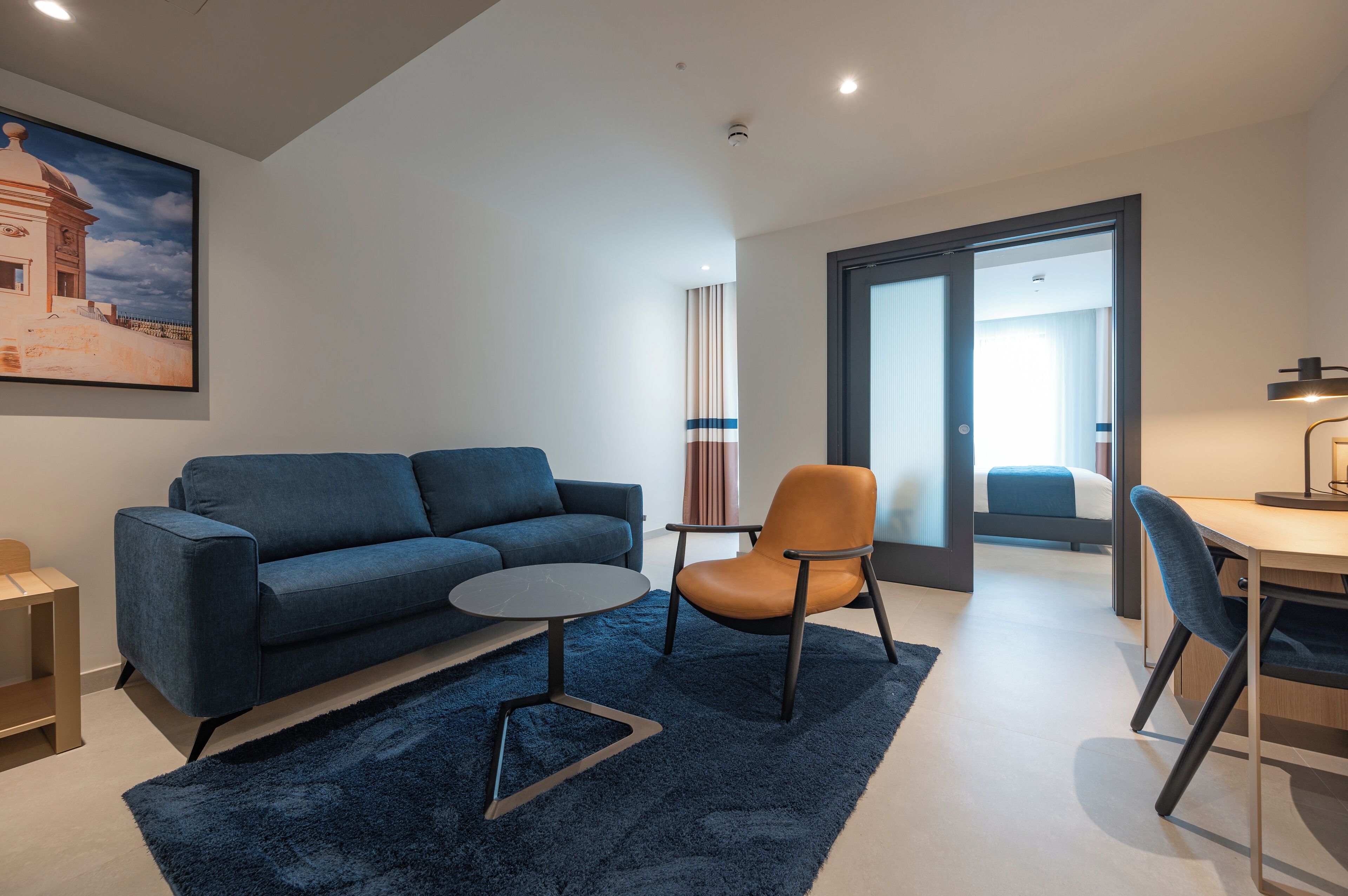 A modern living room with a blue sofa, a round coffee table, and a brown chair. There is a painting on the wall and a door leading to another room.