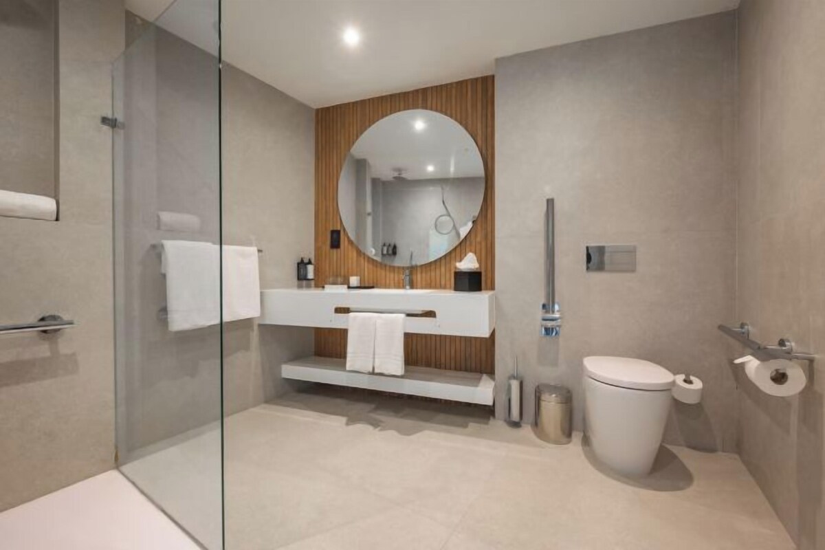 A modern bathroom with a glass shower enclosure, a wooden vanity with a round mirror, a toilet, and a glass shower door.