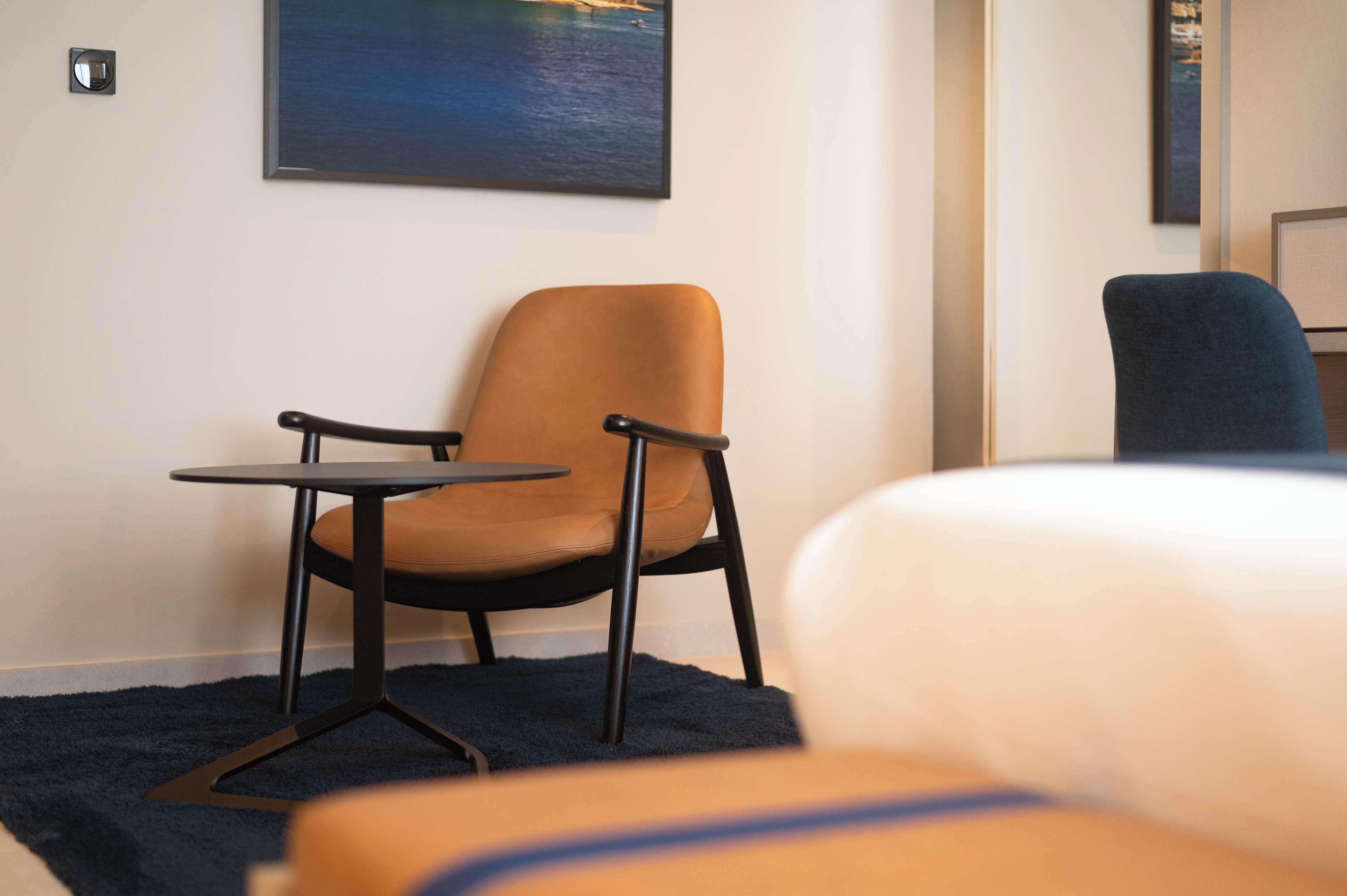 A hotel room with a bed, a brown chair, a small table, and a painting of a landscape on the wall.