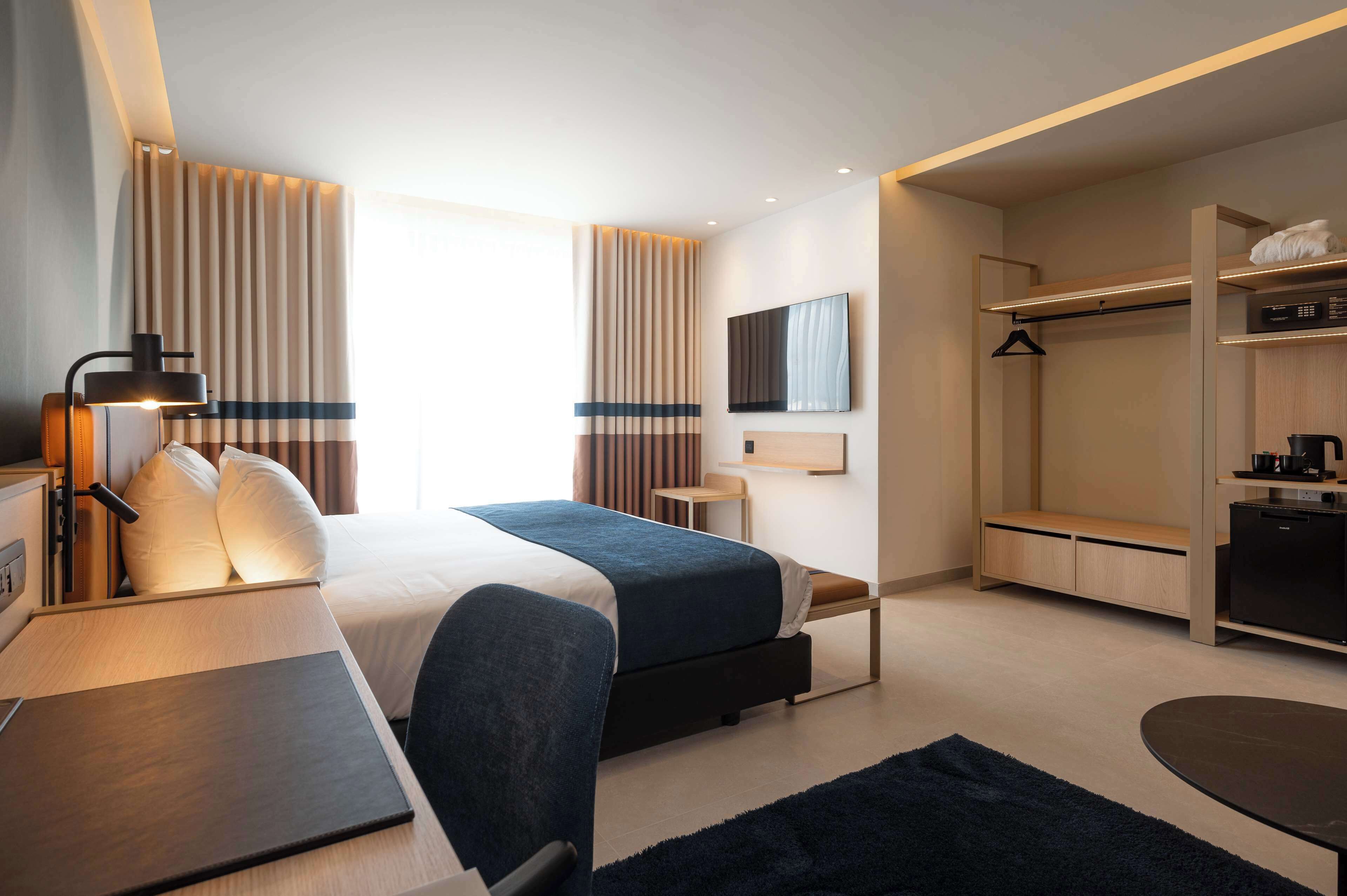 A modern hotel room with a bed, bedside table, lamp, and a view of the city.