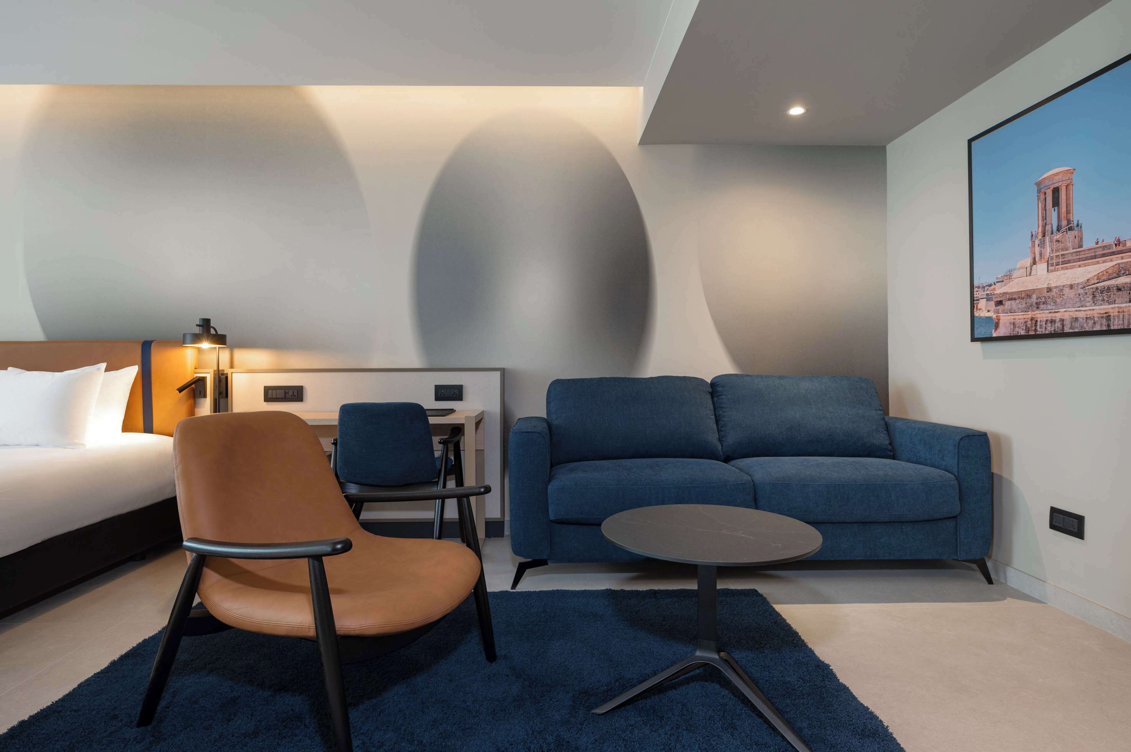 Een moderne hotelkamer met een bed, een blauwe bank, een ronde salontafel en een stoel.
