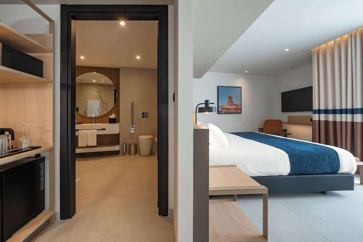 A modern hotel room with a bed, bedside table, and a desk with a chair. There is a bathroom with a mirror, sink, and toilet visible through an open door.