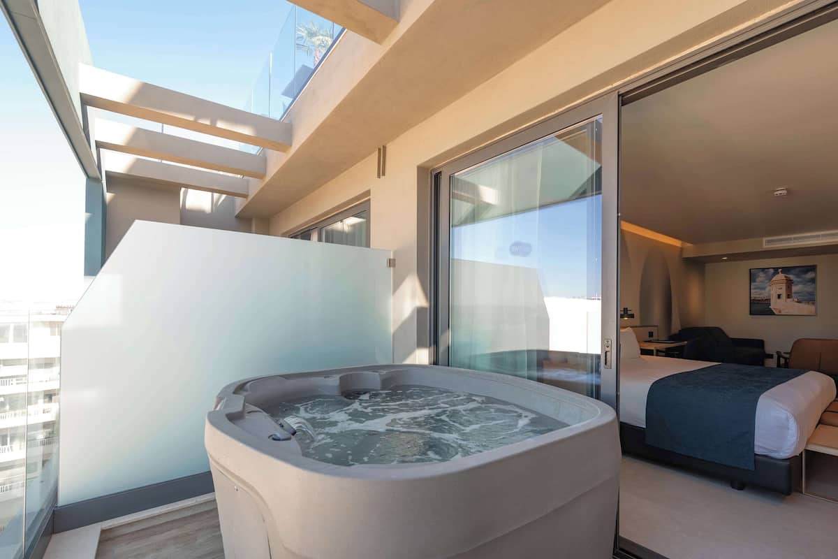 A modern hotel room with a large glass wall, a hot tub, a bed, and a desk.